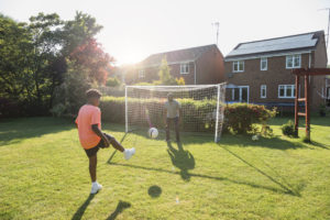 A young boy kicking a football at a goal in his back garden at home. His grandfather is standing in goal, ready to defend it.